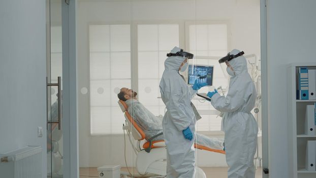 Dental team of dentists consulting patient in cabinet at oral clinic. Caucasian women with dentistry occupation wearing virus protection suits, using stomatology equipment for teeth care