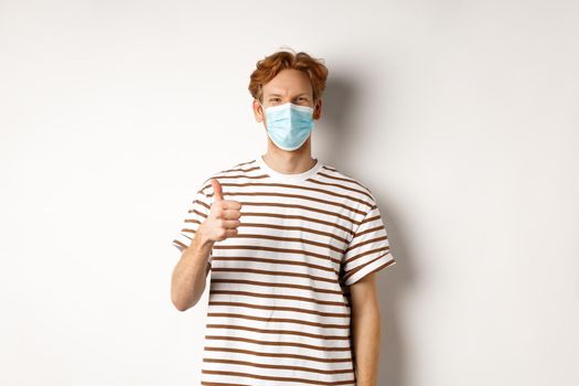 Covid-19, pandemic and social distancing concept. Young man with red hair wearing medical mask to prevent catching coronavirus, showing thumbs-up, white background.