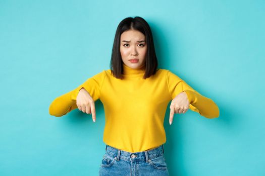 Shopping concept. Sad and worried asian girl frowning silly, pointing fingers down at mistake, standing upset over blue background.