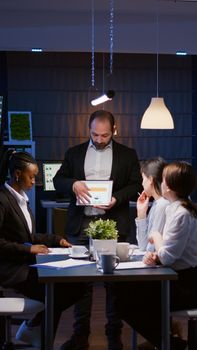 Workaholic overworked businessman explaining company strategy using tablet for presentation working overtime in meeting office room late at night. Diverse multi-ethnic teamwork brainstorming ideas