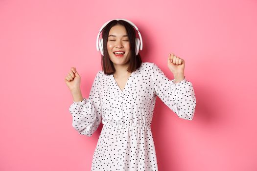 Happy young asian woman dancing and having fun, listening music in headphones, standing over pink background. Copy space