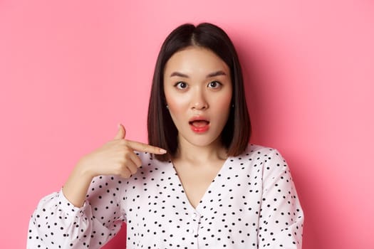 Image of confused asian woman pointing at herself, drop jaw and staring questioned at camera, standing over pink background.