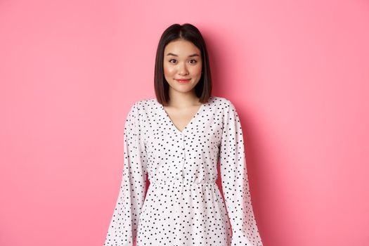 Beautiful asian lady smiling at camera, wearing cute romantic dress, standing over pink background. Copy space