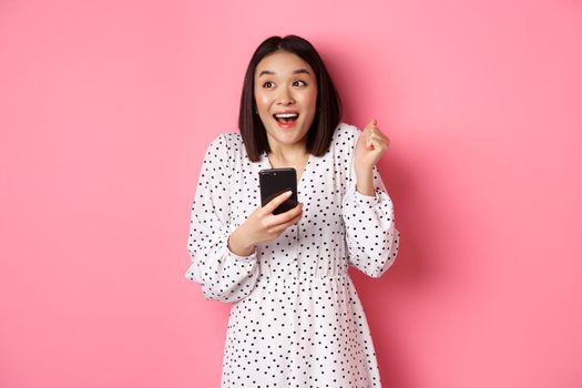 Online shopping and beauty concept. Excited asian woman winning in internet, holding smartphone and rejoicing, smiling happy and celebrating, standing over pink background.