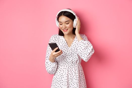 Cute asian girl listening music on headphones, looking at mobile phone and smiling, standing in dress against pink background.
