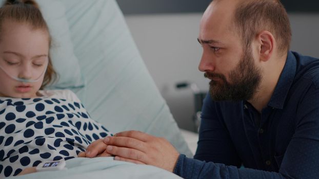 Worried emotional father crying beside sick daughter waiting for healthcare treatment during sickness recovery consultation in hospital ward. Child sleeping recovering after medical surgery