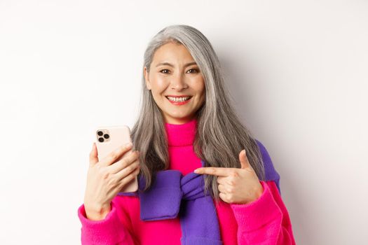 Close up of pretty asian senior woman with grey hair, pointing finger at smartphone and smiling, recommending mobile app, standing over white background.