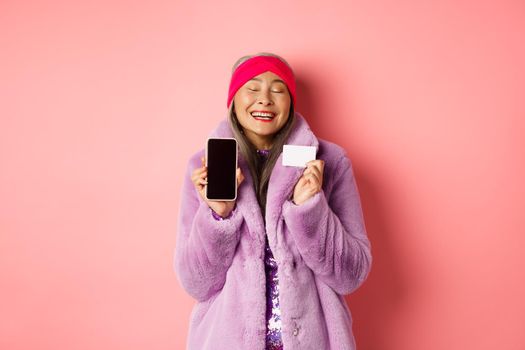 Special promotion. Excited asian senior woman showing blank smartphone screen and plastic credit card, checking out online offer, pink background.