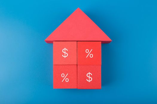 Red house made of toy cubes with percentage sign and dollar on blue background. Modern concept of inflation, percent, mortgage, interest rate growth and increasing