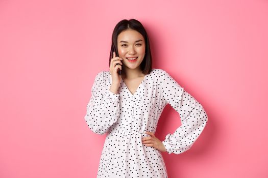 Beautiful asian woman having mobile conversation, making phone call and smiling, standing over pink background.