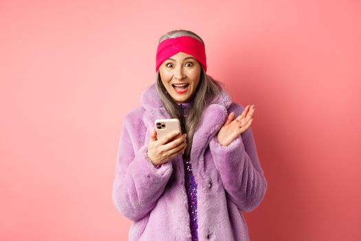 Online shopping and fashion concept. Asian mature woman looking excited as checking out internet promo offer on smartphone, looking happy at camera, pink background.