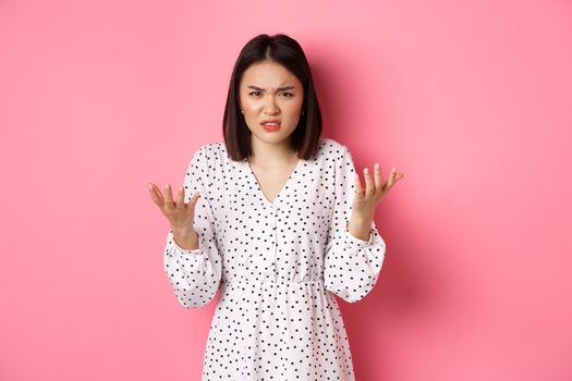 Angry and confused asian woman pointing hands at camera and grimacing furious, standing annoyed against pink background.