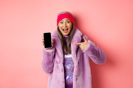 Online shopping and fashion concept. Stylish asian senior woman in faux fur coat pointing finger at blank smartphone screen, smiling happy, showing application on phone.