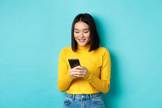 Attractive asian woman reading smartphone screen and smiling, social networking with mobile phone, standing over blue background.
