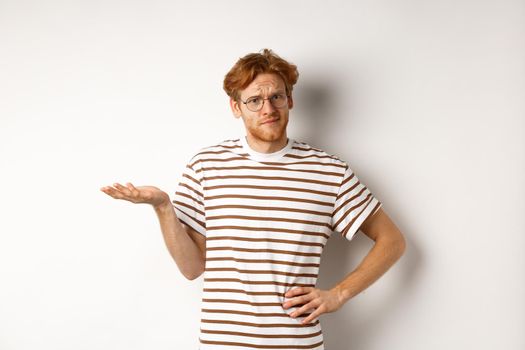 So what. Confused and annoyed redhead man waiting for reply, standing questioned with raised hand, holding something on palm, white background.