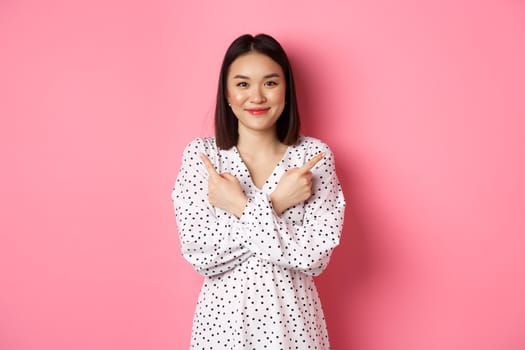 Beautiful asian girl in romantic dress pointing fingers sideways, showing two variants on shopping, smiling at camera, standing against pink background.