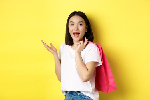 Cheerful asian female shopper looking amused, holding shopping bag and standing over yellow background.
