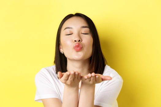 Valentines day concept. Close up of beautiful asian woman pucker lips and holding hands near mouth, blow air kiss at camera with closed eyes, standing over yellow background.