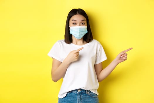 Covid-19, pandemic and social distancing concept. Surprised and happy asian woman in medical mask, showing advertisement, pointing right and smiling, yellow background.