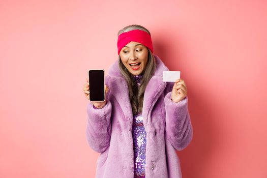 Special promotion. Excited asian senior woman showing blank smartphone screen and plastic credit card, checking out online offer, pink background.