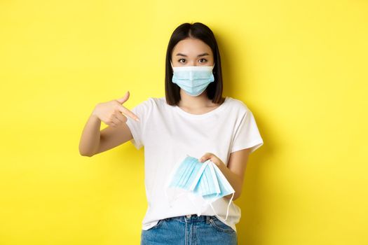 Coronavirus, quarantine and medicine concept. Young asian woman pointing finger at medical masks for going outdoors, standing over yellow background.