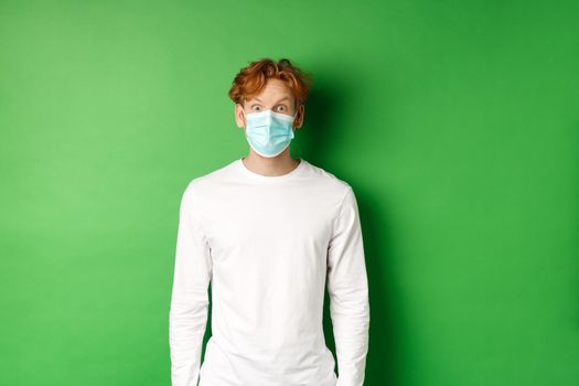 Covid-19, social distancing and lifestyle concept. Young redhead man wearing face mask during coronavirus pandemic, looking surprised at camera, green background.