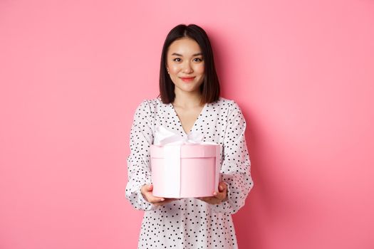Beautiful asian woman wishing happy holidays, giving you gift in cute box, standing against pink background.