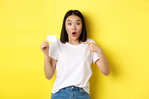 Young asian woman in casual white t-shirt showing plastic credit card and smiling at camera, yellow background.