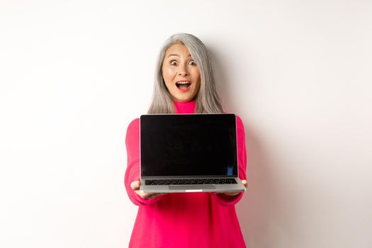 E-commerce concept. Amazed asian senior woman showing blank laptop screen and looking happy, demonstrating promo, white background.