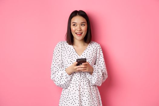 Cute asian woman thinking and smiling, looking left dreamy while messaging on smartphone, browing online stores, standing over pink background.