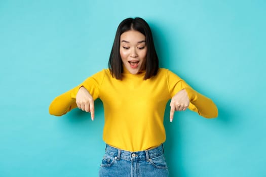 Shopping concept. Surprised cute asian girl checking out discounts, pointing fingers down and looking amazed, saying wow, standing over blue background.