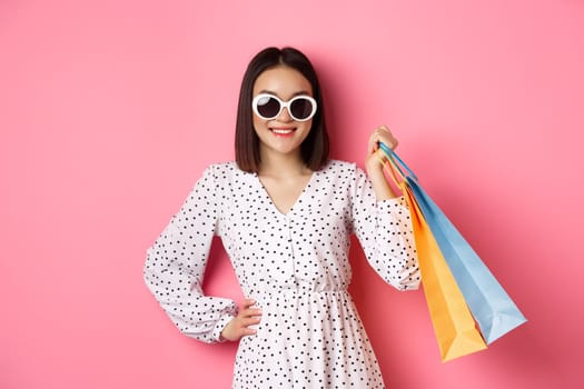 Attractive smiling asian woman holding shopping bags, wearing sunglasses and cute dress, standing against pink background.