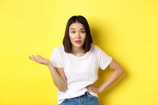 Careless asian woman asking so what, raise hand up and staring skeptical at camera, standing over yellow background.