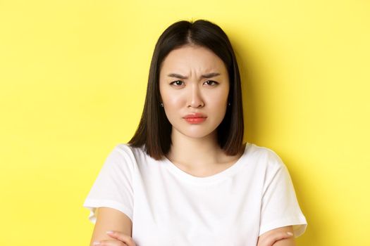 Close up of young asian woman looking angry or disappointed, frowning displeased at camera, standing in white t-shirt, yellow background.