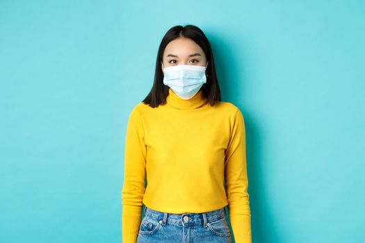 Covid-19, social distancing and pandemic concept. Teenage asian woman in medical mask, looking at camera and standing against blue background in yellow sweater.