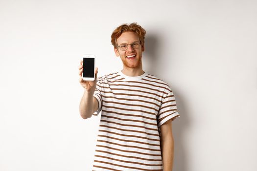 Technology and e-commerce concept. Happy young redhead man in glasses showing blank smartphone screen, smiling satisfied, standing over white background.