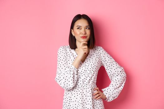 Thoughtful happy asian woman making decision, smiling satisfied and looking at upper left logo, thinking or choosing, standing in dress against pink background.