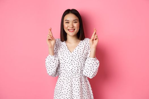 Cute asian woman making a wish, smiling happy and cross fingers for good luck, waiting over pink background.