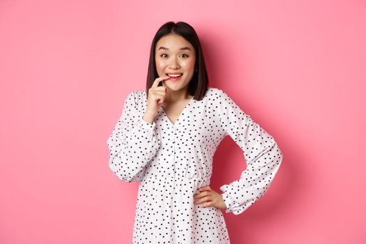 Image of beautiful young lady in dress, biting finger and smiling, staring with temptation and amazement, want buy something, standing over pink background.