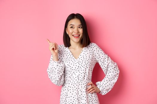 Happy asian girl in dress smiling satisfied, pointing finger at upper left corner, nod in approval, standing in dress against pink background.
