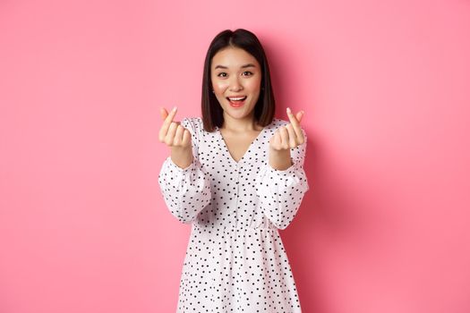 Lovely asian woman in dress showing korean heart signs and smiling, standing on romantic pink background.