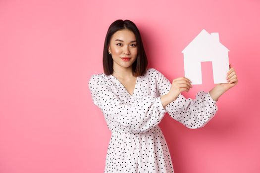 Real estate. Beautiful asian woman demonstrating paper house model, looking at camera confident, advertising home for sale, standing over pink background.