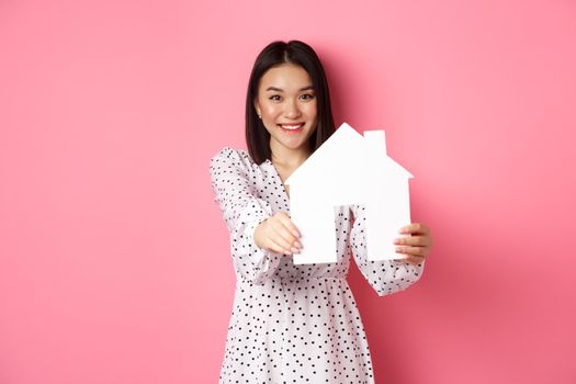 Real estate. Adult asian woman searching for home, holding house model and smiling, promo of broker company, standing over pink background.