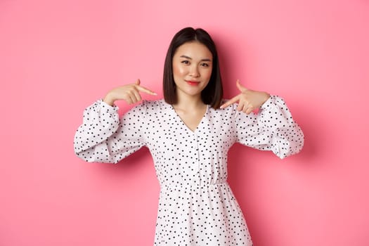 Beautiful asian woman looking confident, pointing at herself and smiling, showing logo on chest, standing over pink background.