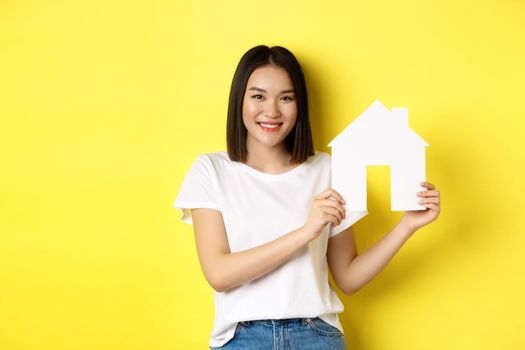 Real estate concept. Smiling beautiful woman showing paper house cutout and looking at camera, buying property, standing over yellow background.