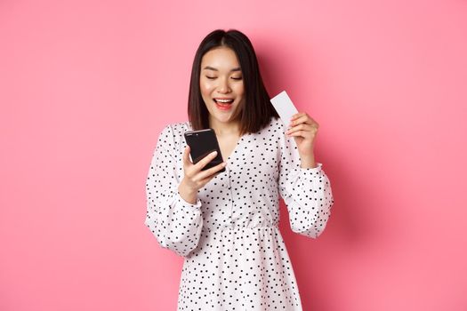 Attractive young asian woman order online, holding credit card and mobile phone, making internet purchase, standing happy over pink background.