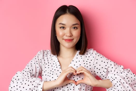 Beauty and lifestyle concept. Close-up of lovely asian woman showing heart sign, smiling and feeling romantic on valentines day, standing over pink background.