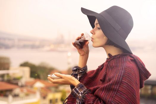 A young girl in a poncho and a hat drinks fragrant delicious tea on the background of the ocean while traveling.