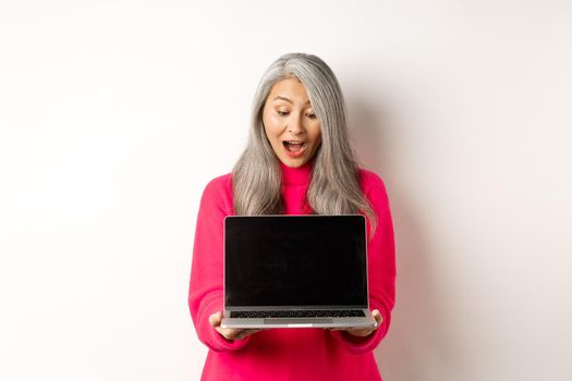 E-commerce concept. Amazed asian grandmother with grey hair, checking out promo online, showing laptop black screen, standing over white background.