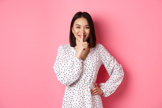Beautiful Korean woman in trendy dress asking to keep secret, hushing with soft smile, planning a surprise, standing over pink background.